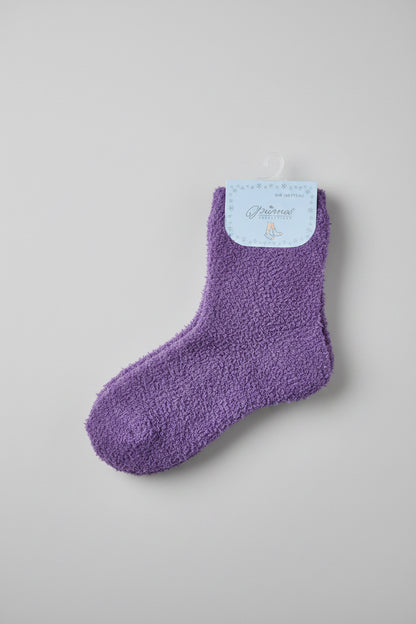 A pair of plush purple socks hanging from a hanger on a dark surface, with a person's feet in sandals and blue jeans visible at the bottom edge.