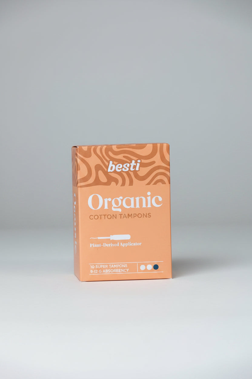 A package of Besti Organic Cotton Tampons on a dark fabric background. The box is a warm peach-orange color with brown wavy patterns at the top and white text.